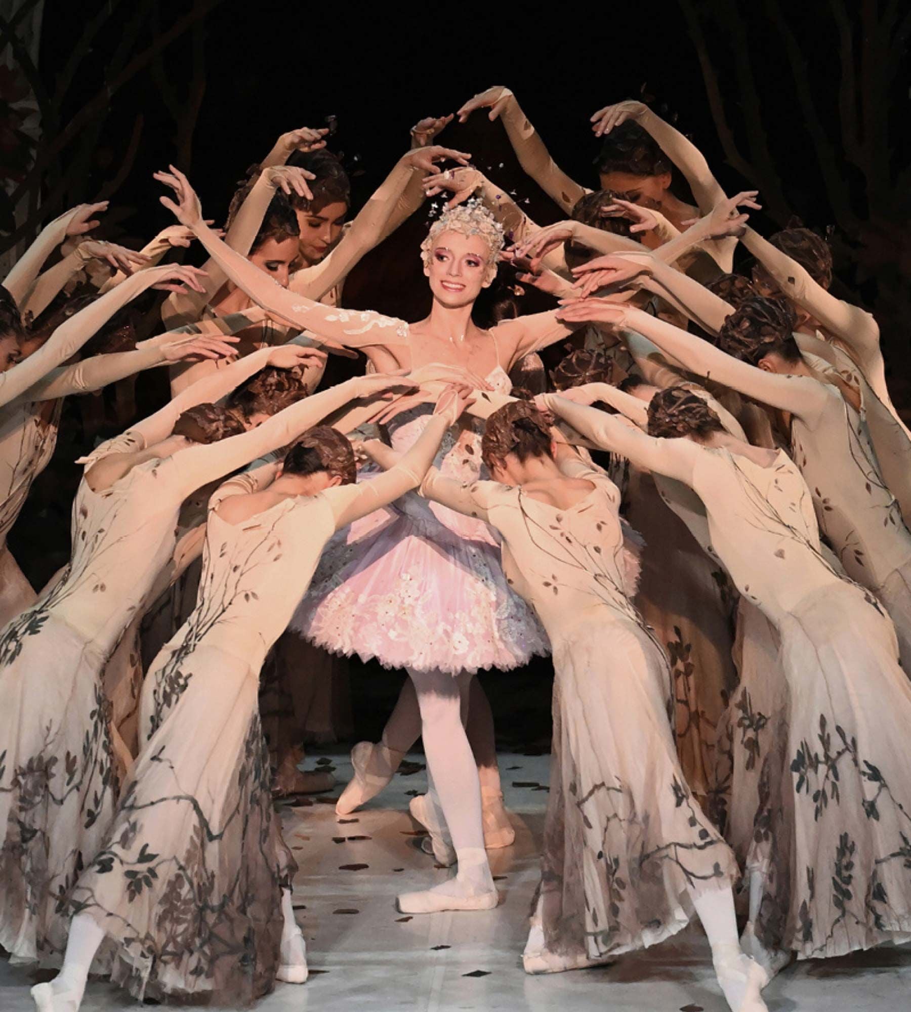 Prima ballerina in pink tutu encircled by dancers in white dresses with raised hands.