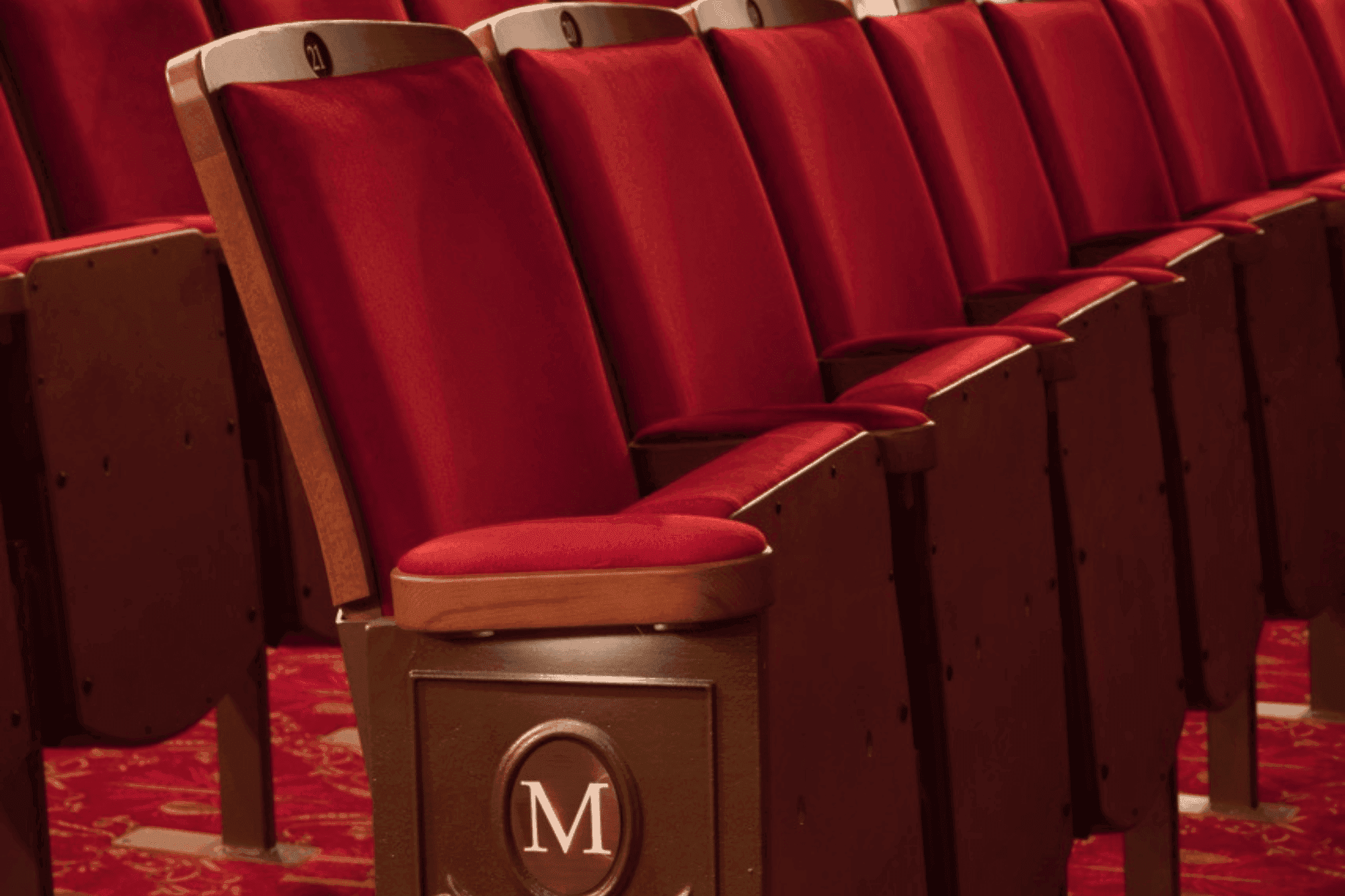 Empty red velvet theater seats with wooden armrests, one displaying an 'M' logo.