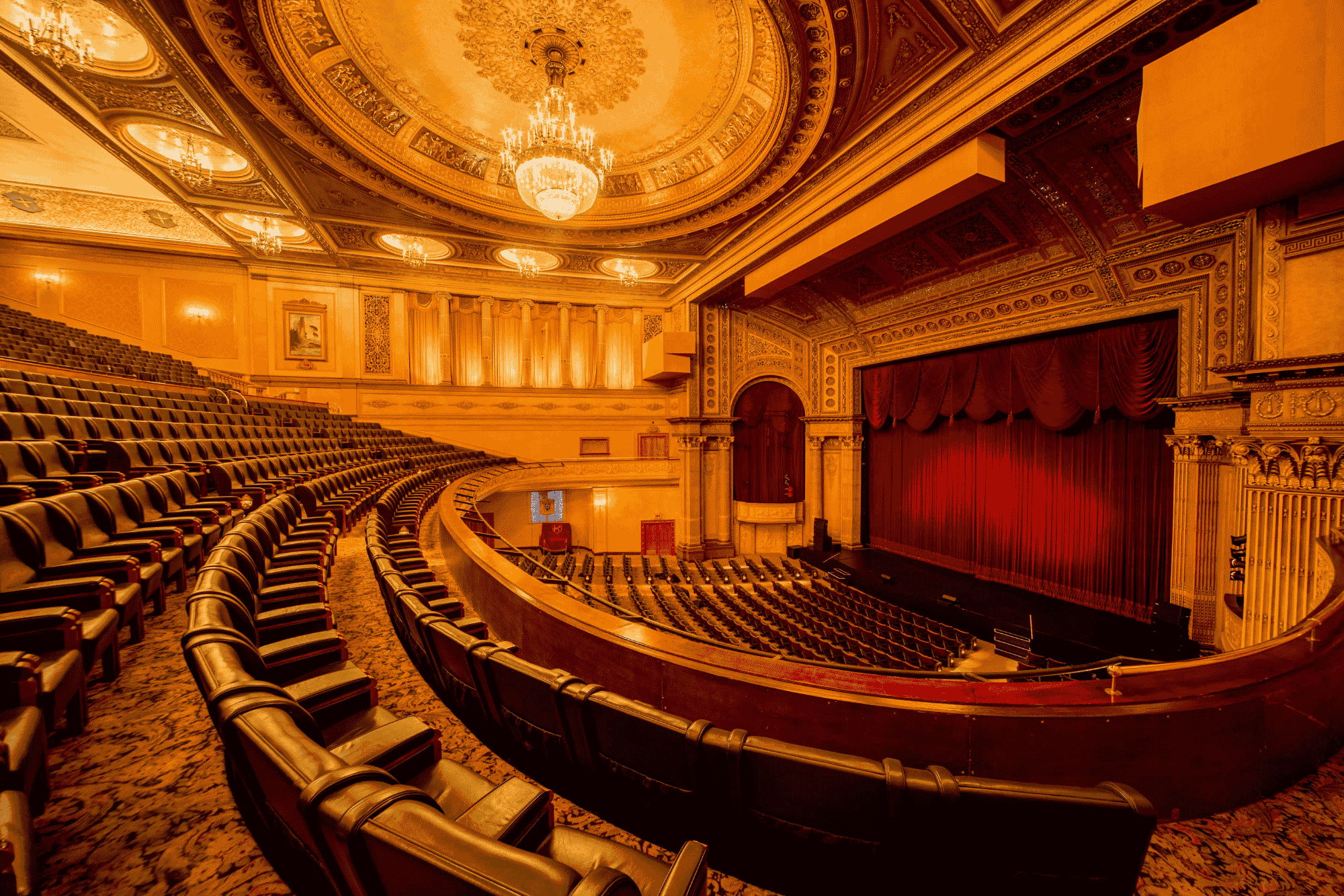 Grand theater interior with ornate ceiling, chandelier, empty seats, and a stage with red curtains.