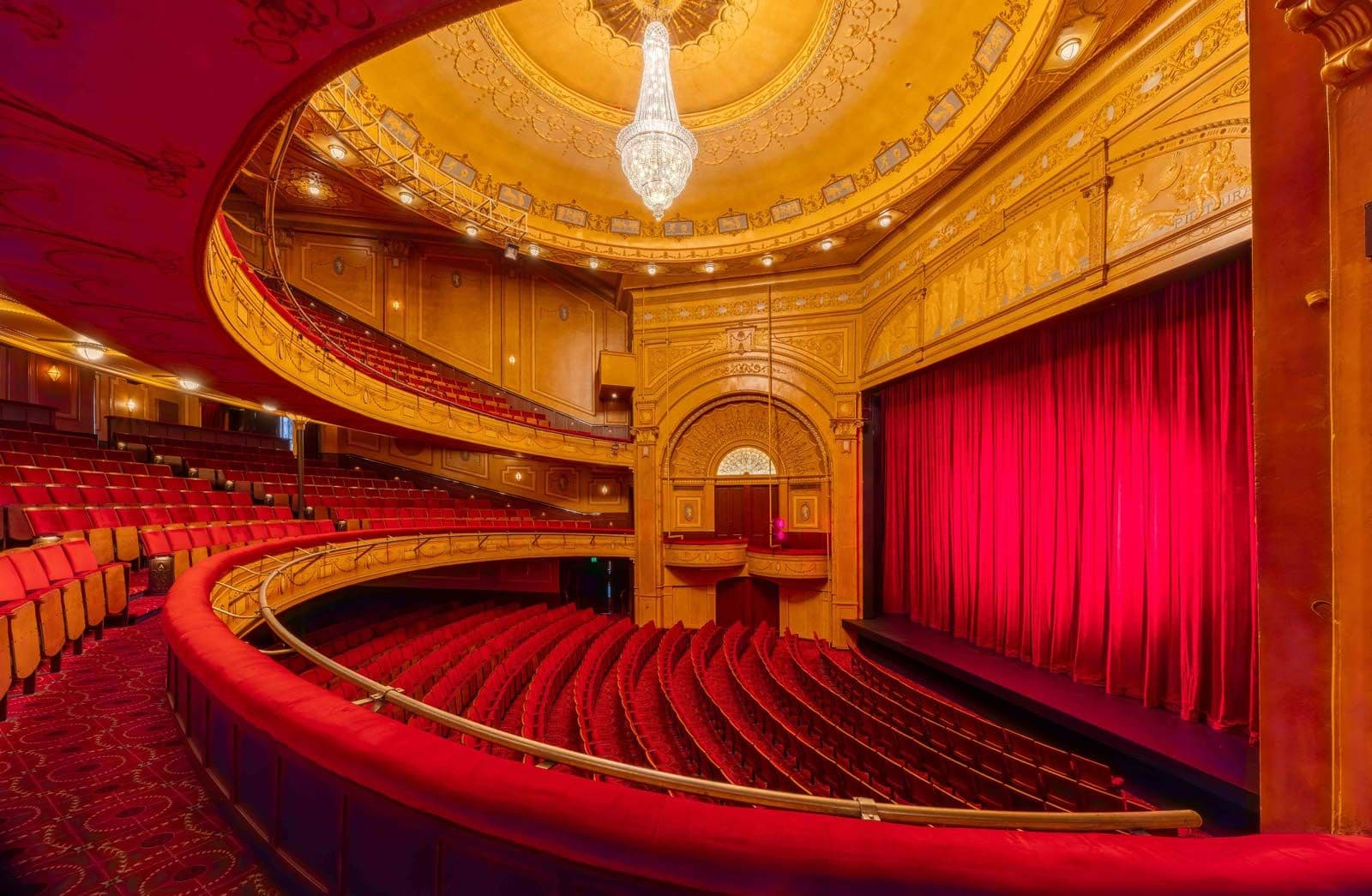 Luxurious theater interior with red velvet seats, grand stage, and red curtain.