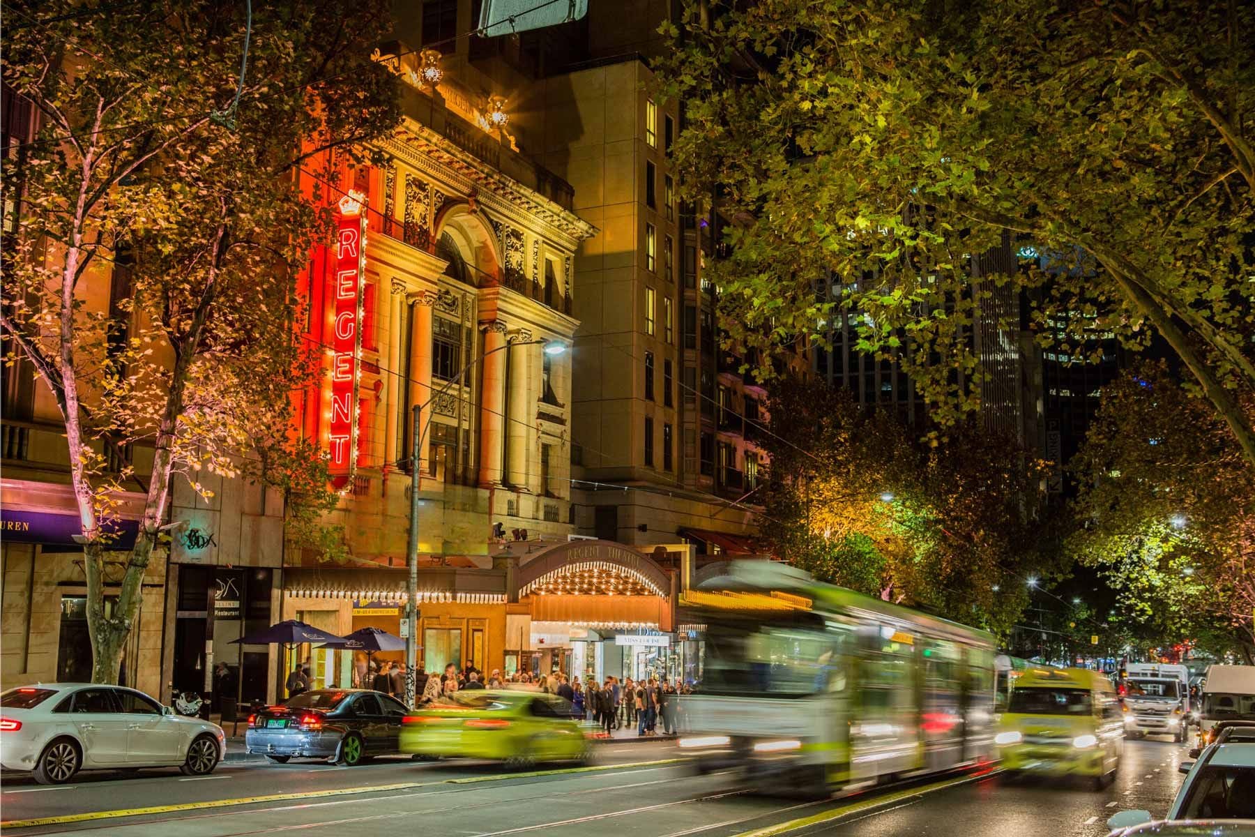 Bustling city street at night with illuminated building, blurred cars and tram.